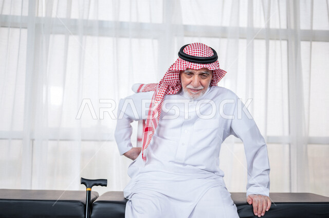 Portrait of an elderly Saudi Arabian Gulf man with his hand on his back, feeling a state of pain, close-up of an elderly Saudi man wearing a white dress with a shemagh and headband, the concept of facial expressions and body language, white background.