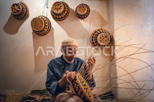 حرفة يدوية قديمة تاريخية, مهنة شعبية تقليدية, منتج صناعي محلي وطني، رجل عربي خليجي سعودي يجلس على الأرض يقوم بجدل سعائف الخوص، استخدام أوراق النخيل المجففة في صناعة السلل, الأصالة والتراث في المملكة العربية السعودية, مهرجان الجنادرية التراثي الثقافي في الرياض 