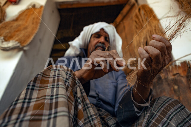 استخدام أوراق النخيل المجففة في صناعة السلل, الأصالة والتراث في المملكة العربية السعودية, حرفة يدوية قديمة تاريخية, مهنة شعبية تقليدية, منتج صناعي محلي وطني، رجل عربي خليجي سعودي يجلس على الأرض يقوم بجدل سعائف الخوص، مهرجان الجنادرية التراثي الثقافي في الرياض