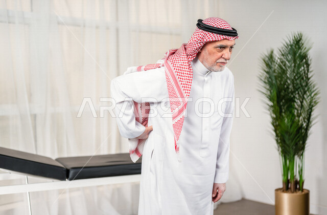 Portrait of an elderly Saudi Arabian Gulf man with his hand on his back, feeling a state of pain, close-up of an elderly Saudi man wearing a white dress with a shemagh and headband, the concept of facial expressions and body language, white background.