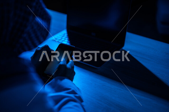 صورة مقربة ليد رجل عربي خليجي سعودي يستخدم الهاتف المحمول ويجلس امام الحاسوب المحمول، يقوم باتصال فيديو ،نقل البيانات والملفات بين الجهازين،  التسويق للتطبيقات الالكترونية ، التعليم الالكتروني، مشاهدة الافلام والفيديو