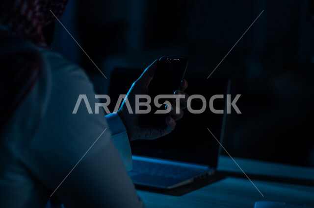 صورة مقربة ليد رجل عربي خليجي سعودي يستخدم الهاتف المحمول ويجلس امام الحاسوب المحمول، يقوم باتصال فيديو ،نقل البيانات والملفات بين الجهازين،  التسويق للتطبيقات الالكترونية ، التعليم الالكتروني، مشاهدة الافلام والفيديو
