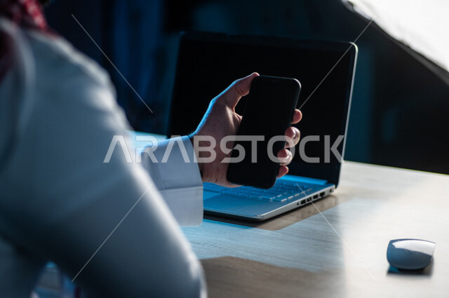 A close-up of the hand of a Saudi Arabian Gulf man using a mobile phone and sitting in front of a laptop, making video calls, transferring data and files between the two devices, marketing for electronic applications, e-learning, watching movies and video