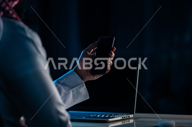 صورة مقربة ليد رجل عربي خليجي سعودي يستخدم الهاتف المحمول ويجلس امام الحاسوب المحمول، يقوم باتصال فيديو ،نقل البيانات والملفات بين الجهازين،  التسويق للتطبيقات الالكترونية ، التعليم الالكتروني، مشاهدة الافلام والفيديو