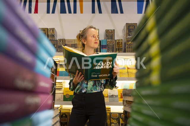 قضاء أوقات ممتعة في شراء الروايات المفيدة للتعلم والتثقف، سائحة أجنبية تمسك بيدها مجلد تعريفي عن المملكة في معرض الكتاب الدولي في الرياض، معلم ومكان سياحي ثقافي مشهور، معارض وفعاليات في السعودية، استقطاب وجذب السياح من أنحاء العالم