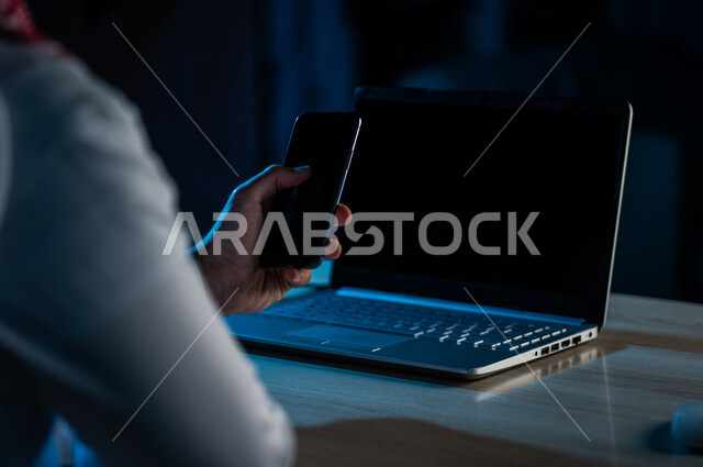 صورة مقربة ليد رجل عربي خليجي سعودي يستخدم الهاتف المحمول ويجلس امام الحاسوب المحمول، يقوم باتصال فيديو ،نقل البيانات والملفات بين الجهازين،  التسويق للتطبيقات الالكترونية ، التعليم الالكتروني، مشاهدة الافلام والفيديو
