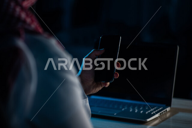 A close-up of the hand of a Saudi Arabian Gulf man using a mobile phone and sitting in front of a laptop, making video calls, transferring data and files between the two devices, marketing for electronic applications, e-learning, watching movies and video