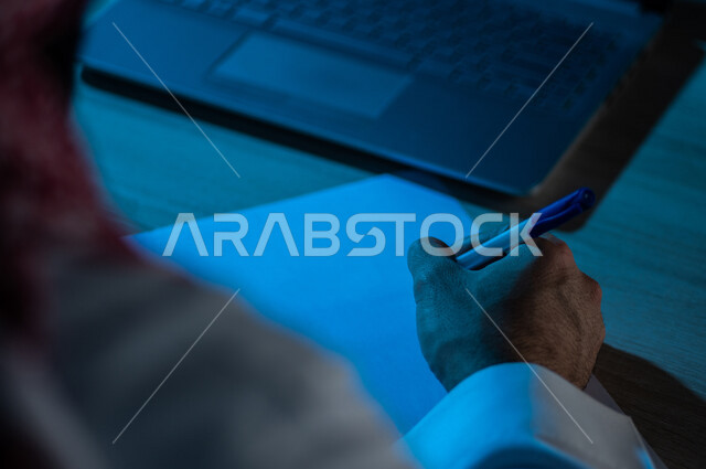صورة مقربة لرجل عربي خليجي سعودي يجلس امام الحاسوب المحمول ، يقوم بتدوين الملاحظات الخاصة بالعمل ، اعداد التقارير ومهام العمل ، تعليم الكتروني ، بحث علمي