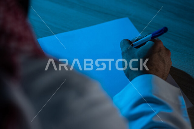 صورة مقربة لرجل عربي خليجي يستخدم الورقة والقلم، يقوم بتدوين الملاحظات الخاصة بالعمل ، اعداد التقارير ومهام العمل ، بحث علمي ، كتابة وتاليف القصص والروايات