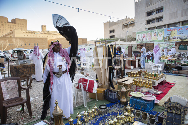 سوق شعبي لبيع الاواني والادوات الشعبيه، بائع عربي خليجي سعودي يرتدي الثوب التقليدي والشماغ والبشت يقف في بازار سياحي تراثي، الأصالة والتراث في المملكة العربية السعودية, منتج صناعي محلي وطني