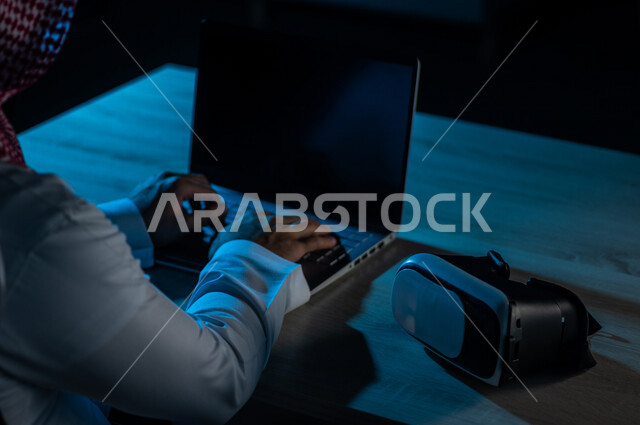 صورة مقربة لرجل عربي خليجي سعودي يستخدم الحاسوب المحمول وبجانبه نظارة الواقع الافتراضي ، ربط التعليم بالواقع الافتراضي ، التسلية والالعاب، نظارة ثلاثية الابعاد تحاكي الواقع الافتراضي