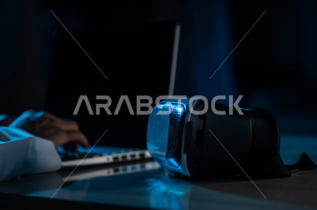 صورة مقربة لرجل عربي خليجي سعودي يستخدم الحاسوب المحمول وبجانبه نظارة الواقع الافتراضي ، ربط التعليم بالواقع الافتراضي ، التسلية والالعاب، نظارة ثلاثية الابعاد تحاكي الواقع الافتراضي