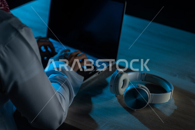 صورة مقربة لرجل عربي خليجي سعودي يستخدم الحاسوب المحمول وبجانبه سماعة الراس لاستخدامها في التعليم عن بعد ، الدورات الالكترونية ، اجتماع اونلاين ، الاستماع الى الموسيقى
