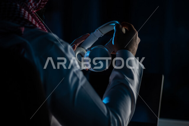 صورة مقربة لرجل عربي خليجي سعودي يحمل بيده سماعة الراس لاستخدامها في التعليم عن بعد ، الدورات الالكترونية ، اجتماع اونلاين ، الاستماع الى الموسيقى