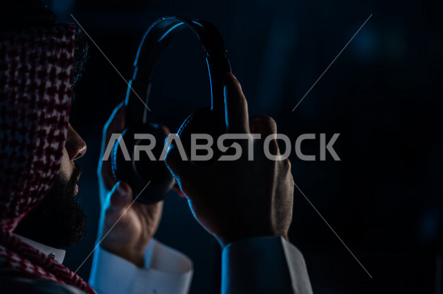 صورة مقربة لرجل عربي خليجي سعودي يحمل بيده سماعة الراس لاستخدامها في التعليم عن بعد ، الدورات الالكترونية ، اجتماع اونلاين ، الاستماع الى الموسيقى