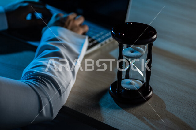 صورة مقربة لساعة رملية بالقرب من رجل عربي خليجي سعودي يستخدم الحاسوب المحمول ، تحديد موعد العمل ، انجاز المهام بدقة وكفاءة ، ادارة الجدول الزمني ، مفهوم ادارة الوقت
