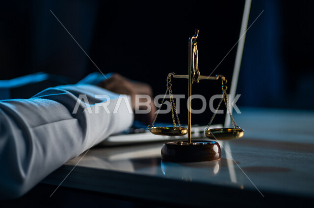 صورة مقربة لميزان العدل بالقرب من رجل عربي خليجي سعودي يستخدم الحاسوب المحمول ، محامي ينجز المهام القانونية ، مفهوم العدالة والقانون