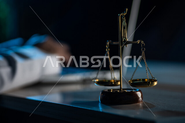 صورة مقربة لميزان العدل بالقرب من رجل عربي خليجي سعودي يستخدم الحاسوب المحمول ، محامي ينجز المهام القانونية ، مفهوم العدالة والقانون
