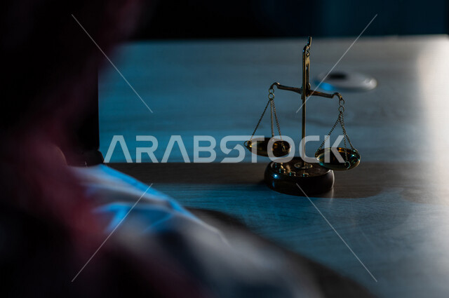 صورة مقربة لميزان العدل بالقرب من رجل عربي خليجي سعودي يستخدم الحاسوب المحمول ، محامي ينجز المهام القانونية ، مفهوم العدالة والقانون