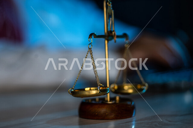 صورة مقربة لميزان العدل بالقرب من رجل عربي خليجي سعودي يستخدم الحاسوب المحمول ، محامي ينجز المهام القانونية ، مفهوم العدالة والقانون