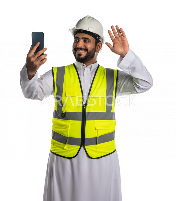 بورتريه لمهندس عربي خليجي سعودي يرتدي الخوذة والسترة الخاصة بالعمل ،يستخدم الهاتف المحمول ،اجراء مكالمة فيديو، اتصال عمل ،خلفية بيضاء