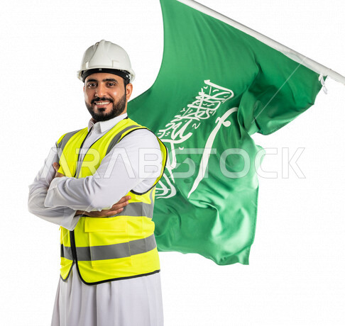 بورتريه لمهندس عربي خليجي سعودي مبتسم وسعيد، يرتدي قبعة و سترة الوقاية ،يقف مكتف اليدين ، خلفه علم المملكة العربية السعودية ، الاحتفال باليوم الوطني السعودي ،العيد الوطني السعودي ، خلفية بيضاء