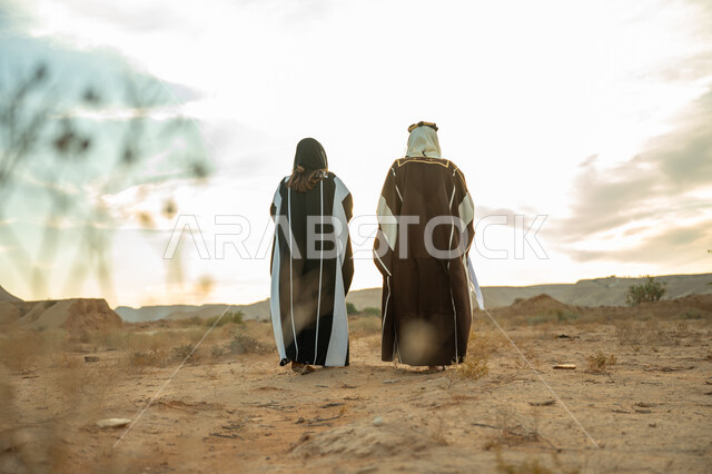 يوم بدينا 22 فبراير, ذكرى تأسيس الدولة السعودية الأولى 1727م, حب الوطن والإنتماء اليه, ارتداء الزي الشعبي في مناسبة وطنية, صورة من الخلف لرجل عربي خليجي سعودي يرتدي البشت والعقال الذهبي وامرأة ترتدي العباءة يسيران على الرمال الذهبية في الصحراء