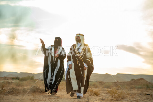 يوم بدينا 22 فبراير, ذكرى تأسيس الدولة السعودية الأولى 1727م, ارتداء الزي الشعبي في مناسبة وطنية, رجل عربي خليجي سعودي يرتدي البشت والعقال الذهبي وامرأة ترتدي العباءة والبرقع يسيران على الرمال الذهبية في الصحراء, الفخر والاعتزاز بالإنتماء الى الوطن