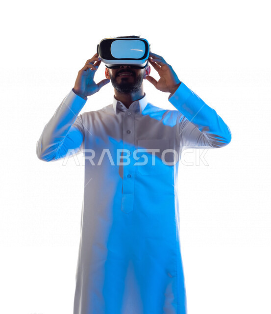 بورتريه لرجل عربي خليجي سعودي مبتسم يرتدي نظارة الواقع الإفتراضي ثلاثية الأبعاد الفريدة و العجيبة ، ويمسكها بيديه ، التفاعل و المتعة و التسلية بمشاهدة الأفلام و محاكاة العالم الإفتراضي ، خلفية بيضاء