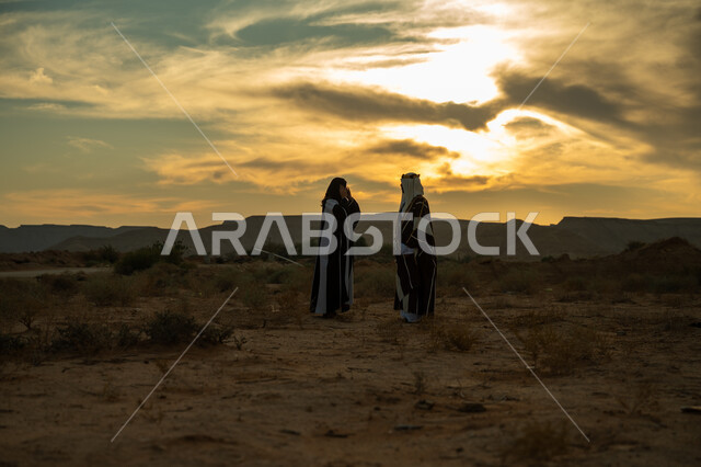 ذكرى تأسيس الدولة السعودية الأولى 1727م, ارتداء الزي الشعبي في مناسبة وطنية, يوم بدينا 22 فبراير, حب الوطن والإنتماء اليه, رجل عربي خليجي سعودي يرتدي البشت والعقال الذهبي وامرأة ترتدي العباءة والبرقع يقفان في الطبيعة الصحراوية وقت الغروب, الطول الكامل للجسم
