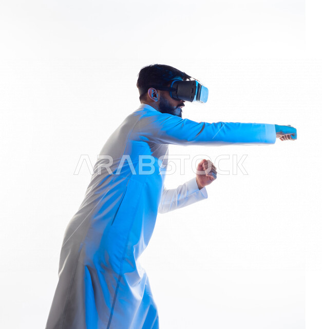 بورتريه لرجل عربي خليجي سعودي يرتدي نظارة الواقع الإفتراضي ثلاثية الأبعاد الفريدة ، يتفاعل بيديه بألعاب الفنون القتالية الدفاعية ، ألعاب العالم الافتراضي ، محاكاة العالم الإفتراضي ، تقنيات و ألعاب ، خلفية بيضاء