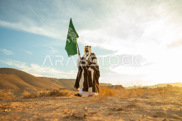 ذكرى تأسيس الدولة السعودية الأولى 1727م, الاحتفال بمناسبة وطنية متميزة, ارتداء زي المنطقة الوسطى, الاعتزاز والفخر بالانتماء للوطن, يوم بدينا 22 فبراير, رجل عربي خليجي سعودي يرتدي البشت والعقال الذهبي يرفع سارية لعلم المملكة في الصحراء في النهار