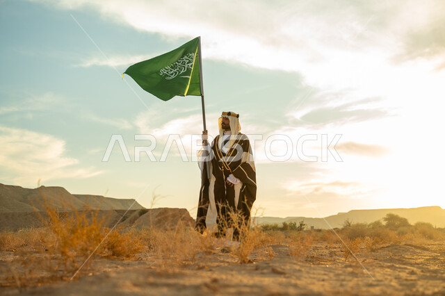 يوم بدينا 22 فبراير, ذكرى تأسيس الدولة السعودية الأولى 1727م, الاحتفال بمناسبة وطنية متميزة, ارتداء زي المنطقة الوسطى, رجل عربي خليجي سعودي يرتدي البشت والعقال الذهبي يرفع سارية لعلم المملكة  في الصحراء في النهار, مفهوم حب الوطن والانتماء اليه