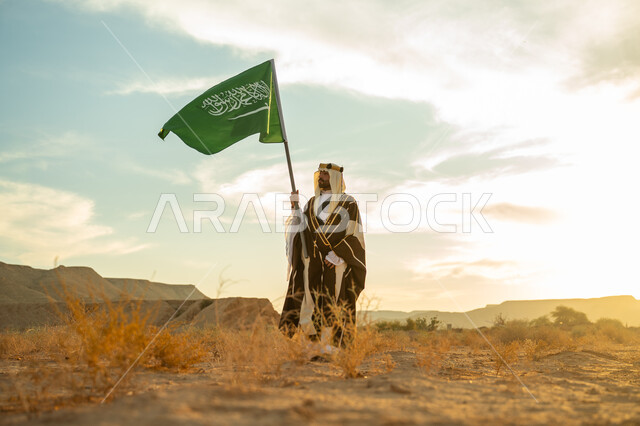 ذكرى تأسيس الدولة السعودية الأولى 1727م, الاحتفال بمناسبة وطنية متميزة, ارتداء زي المنطقة الوسطى, يوم بدينا 22 فبراير, رجل عربي خليجي سعودي يرتدي البشت والعقال الذهبي يرفع سارية لعلم المملكة  في الصحراء في النهار, مفهوم حب الوطن والانتماء اليه