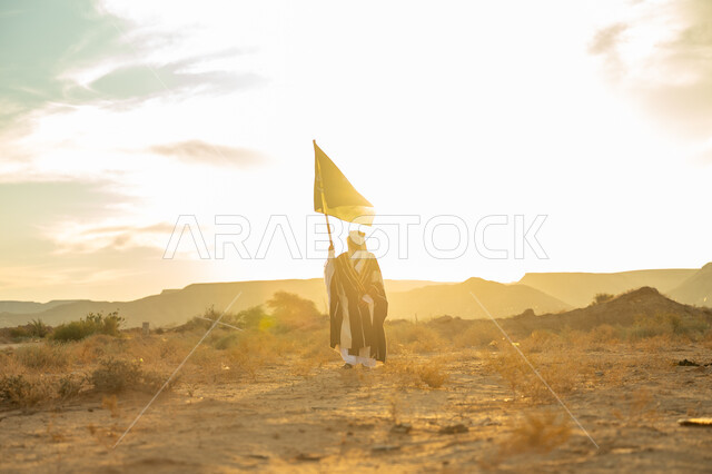 Celebrating a distinguished national occasion, wearing the Central Region costume, the anniversary of the founding of the first Saudi state 1727 AD, the day we started on February 22, a Saudi Gulf Arab man wearing a bisht and a golden headband raises a flagpole for the Kingdom in the desert during the day, the concept of love of the homeland and belonging to it