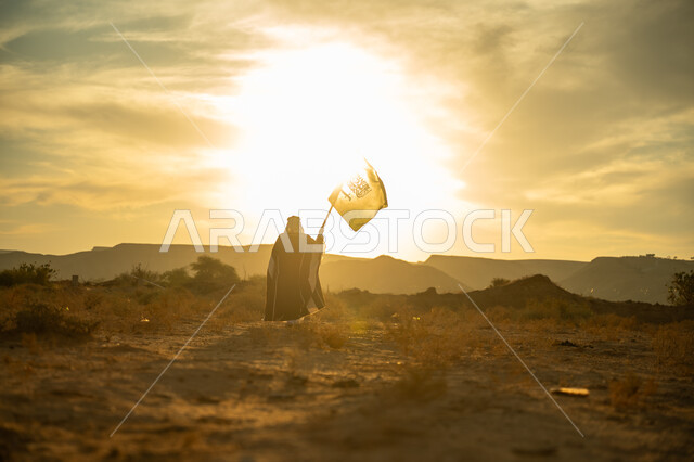 يوم بدينا 22 فبراير, الاعتزاز والفخر بالوطن, ذكرى تأسيس الدولة السعودية الأولى 1727م, الاحتفال بمناسبة وطنية متميزة, ارتداء زي المنطقة الوسطى, صورة من الخلف لرجل عربي خليجي سعودي يرتدي البشت والعقال الذهبي يرفع علم المملكة عند شروق الشمس في الصحراء