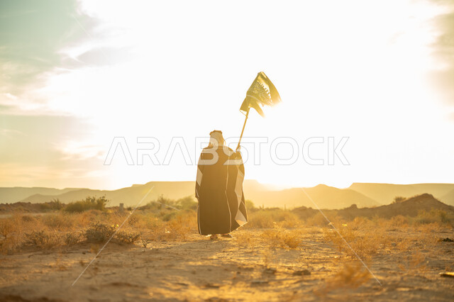 ذكرى تأسيس الدولة السعودية الأولى 1727م, الاحتفال بمناسبة وطنية متميزة, ارتداء زي المنطقة الوسطى, حب الوطن والانتماء اليه, يوم بدينا 22 فبراير, صورة من الخلف لرجل عربي خليجي سعودي يرتدي البشت والعقال الذهبي يرفع علم المملكة عند شروق الشمس في الصحراء