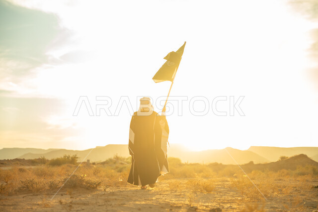 يوم بدينا 22 فبراير, ذكرى تأسيس الدولة السعودية الأولى 1727م, الاحتفال بمناسبة وطنية متميزة, ارتداء زي المنطقة الوسطى, حب الوطن والانتماء اليه, صورة من الخلف لرجل عربي خليجي سعودي يرتدي البشت والعقال الذهبي يرفع علم المملكة عند شروق الشمس في الصحراء