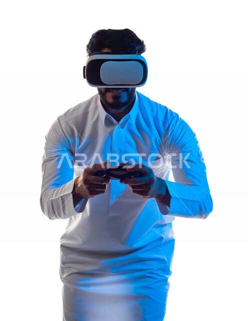 بورتريه لرجل عربي خليجي سعودي يرتدي نظارة الواقع الإفتراضي ثلاثية الأبعاد ، يمسك بيده عصا التحكم و يلعب بألعاب الفيديو بإيماءات تدل على السعادة ،  المتعة و التسلية بألعاب العالم الافتراضي ، ألعاب البلايستيشن الترفيهية ، تقنيات و ألعاب