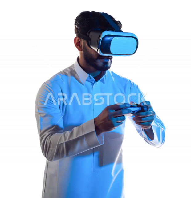 Portrait of a Saudi Arabian Gulf man wearing 3D virtual reality glasses, holding a joystick in his hand and playing video games with gestures that indicate focus, fun and entertainment in virtual world games, PlayStation entertainment games, techniques an