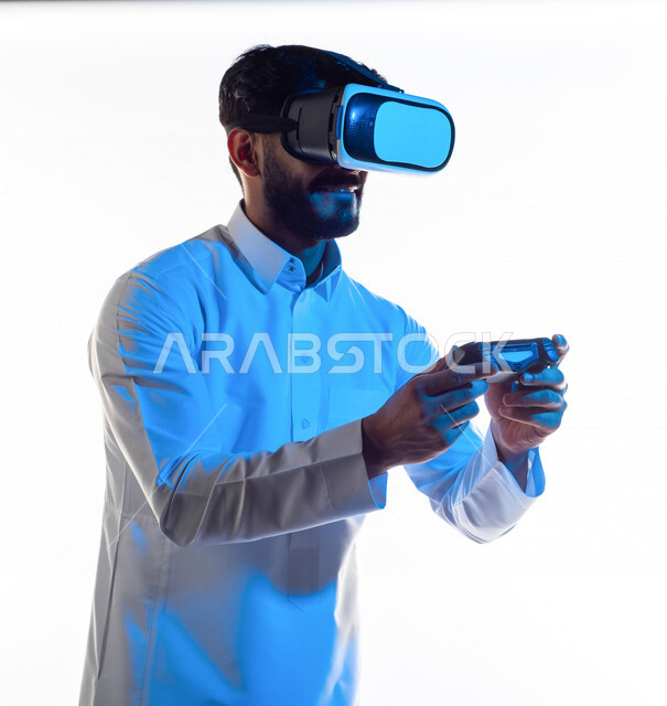 Portrait of a Saudi Arabian Gulf man wearing 3D virtual reality glasses, holding a joystick in his hand and playing video games with gestures indicating happiness, fun and entertainment in virtual world games, PlayStation entertainment games, technologies