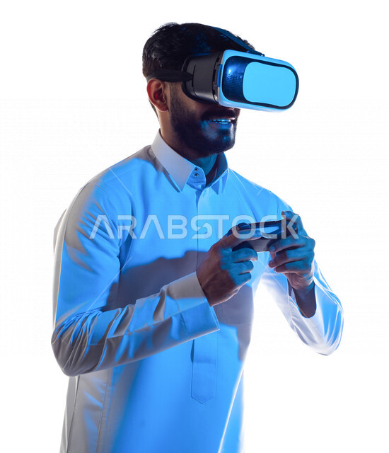 بورتريه لرجل عربي خليجي سعودي يرتدي نظارة الواقع الإفتراضي ثلاثية الأبعاد ، يمسك بيده عصا التحكم و يلعب بألعاب الفيديو بإيماءات تدل على السعادة ،  المتعة و التسلية بألعاب العالم الافتراضي ، ألعاب البلايستيشن الترفيهية ، تقنيات و ألعاب