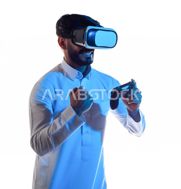 بورتريه لرجل عربي خليجي سعودي يرتدي نظارة الواقع الإفتراضي ثلاثية الأبعاد ، يمسك بيده عصا التحكم و يلعب بألعاب الفيديو بإيماءات تدل على الفوز ،  المتعة و التسلية بألعاب العالم الافتراضي ، ألعاب البلايستيشن الترفيهية ، تقنيات و ألعاب