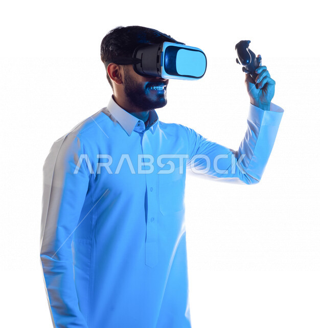 Portrait of a Saudi Arabian Gulf man wearing 3D virtual reality glasses, holding a joystick in his hand and playing video games with gestures indicating victory, fun and entertainment in virtual world games, PlayStation entertainment games, techniques and