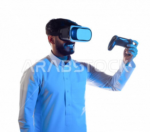 بورتريه لرجل عربي خليجي سعودي يرتدي نظارة الواقع الإفتراضي ثلاثية الأبعاد ، يمسك بيده عصا التحكم و يلعب بألعاب الفيديو بإيماءات تدل على الفوز ،  المتعة و التسلية بألعاب العالم الافتراضي ، ألعاب البلايستيشن الترفيهية ، تقنيات و ألعاب