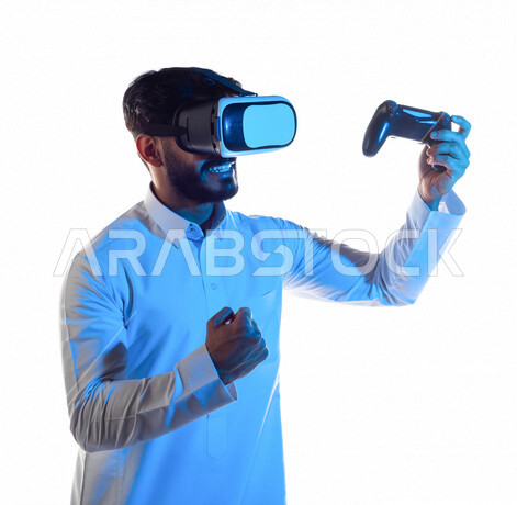 Portrait of a Saudi Arabian Gulf man wearing 3D virtual reality glasses, holding a joystick in his hand and playing video games with gestures indicating victory, fun and entertainment in virtual world games, PlayStation entertainment games, techniques and