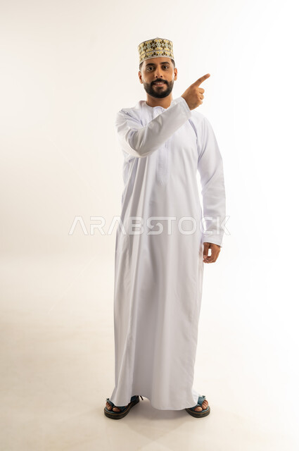 الإشارة لموضع معين, مفهوم الرجولة والأناقة والوسامة, بورتريه لرجل عربي خليجي عماني يرتدي الدشداشة والكمة يشير بإصبع السبابة إلى شيء ما في الأعلى ينظر إلى الكاميرا بإيماءات الثقة بالنفس, الاهتمام بالمظهر الخارجي, الجسم بالطول الكامل, خلفية بيضاء