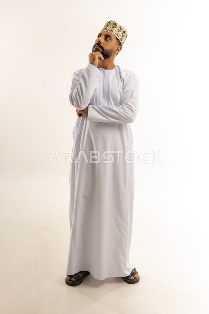 إيماءات محاولة تذكر أمر ما، بورتريه لرجل عربي خليجي عماني يرتدي الدشداشة والكمة يضع يده على ذقنه ينظر إلى الأعلى بتعابير التفكير، التأمل والتخطيط للمستقبل، التركيز والتمعن لإيجاد حل لمشكلة معينة، الجسم بالطول الكامل، خلفية بيضاء