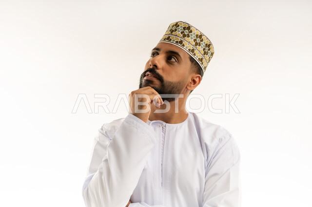 التأمل والتخطيط للمستقبل، إيماءات محاولة تذكر أمر ما، بورتريه مقرب لرجل عربي خليجي عماني يرتدي الدشداشة والكمة يضع يده على ذقنه ينظر إلى الأعلى بتعابير التفكير، التركيز والتمعن لإيجاد حل لمشكلة معينة، خلفية بيضاء