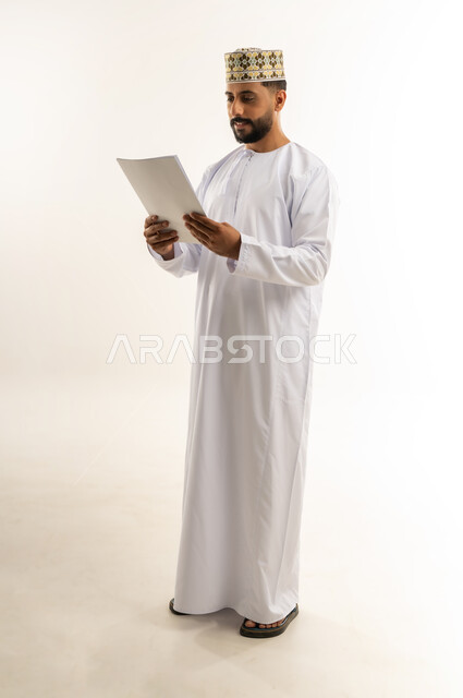 The concept of self-learning, reading, studying and project study, portrait of an Arab Gulf Omani man wearing a dishdasha and a kummah holding a set of papers in his hand and browsing through them with gestures of concentration and contemplation, spending free time in education and learning, full-length body, white background
